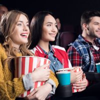 young smiling friends with popcorn watching film in movie theater